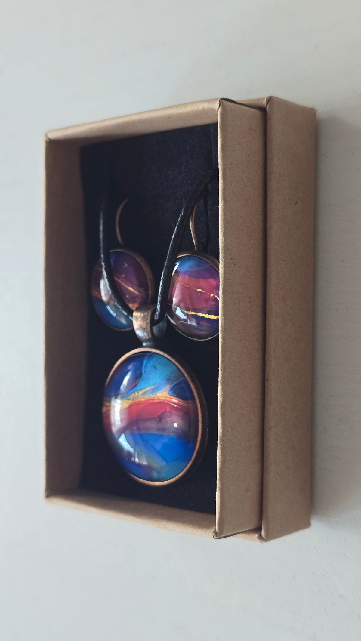 Rundes Cabochon-Schmuckset mit Anhänger und Ohrringen an schwarzem Band, mit abstraktem Muster in Blau, Cyan, Pink und Violett sowie feinen goldenen Linien.