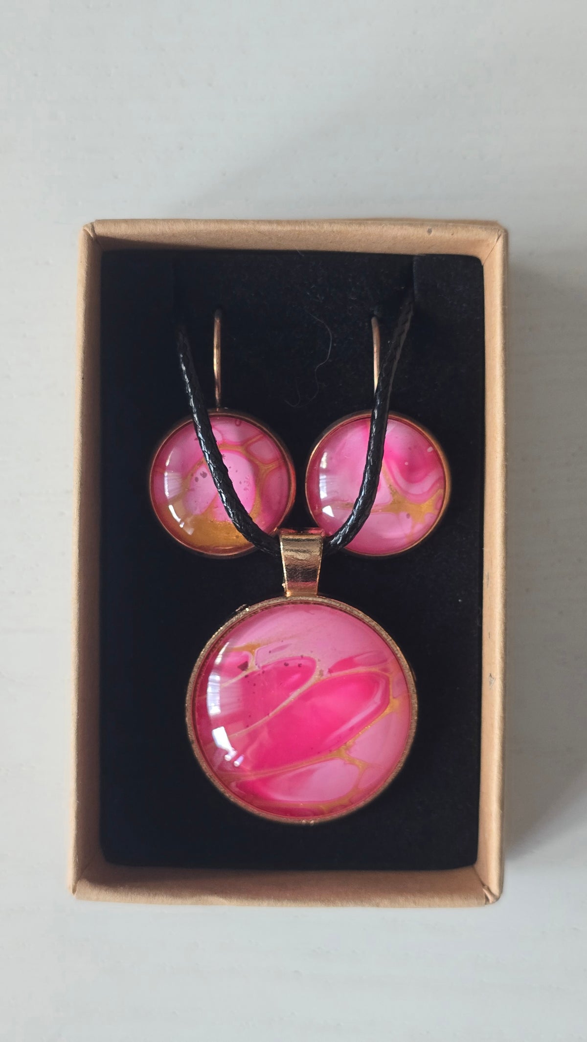 Cabochon-Schmuckset mit Anhänger und Ohrringen in Pink- und Rosatönen mit goldenen, fliessenden Linien und schwarzem Textilband in einer Schachtel.