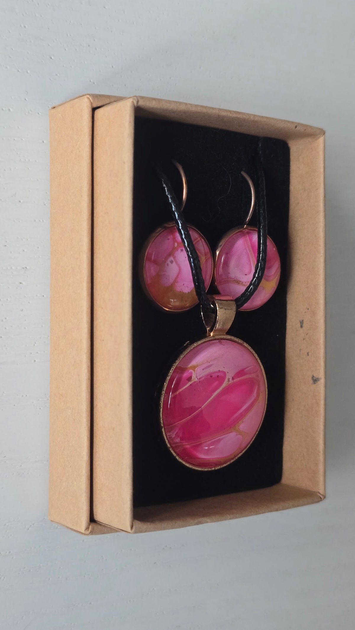 Cabochon-Schmuckset mit Anhänger und Ohrringen in Pink- und Rosatönen mit goldenen, fliessenden Linien und schwarzem Textilband in einer Schachtel.