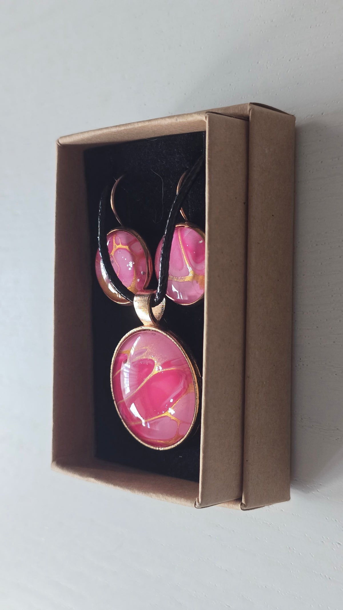 Cabochon-Schmuckset mit Anhänger und Ohrringen in Pink- und Rosatönen mit goldenen, fliessenden Linien und schwarzem Textilband in einer Schachtel.