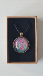 Runder Cabochon-Anhänger an schwarzem Band mit organischem, dunkel umrandetem Muster in Pink, Rosa, Türkis und warm schimmernden Nuancen.