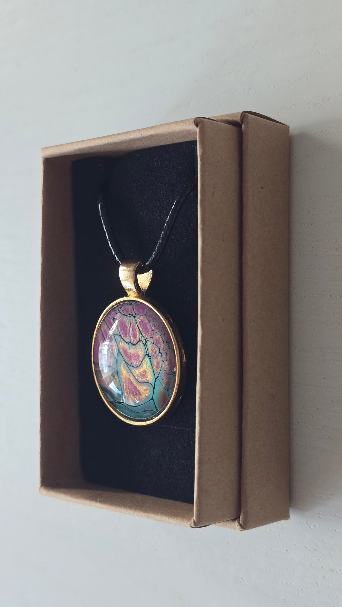 Runder Cabochon-Anhänger an schwarzem Band mit organischem, dunkel umrandetem Muster in Pink, Rosa, Türkis und warm schimmernden Nuancen.