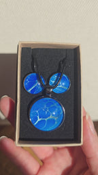 Rundes Cabochon-Schmuckset mit Anhänger und Ohrringen in intensivem Blau und Türkis, durchzogen von feinen silbernen Strukturen, an schwarzem geflochtenem Band.