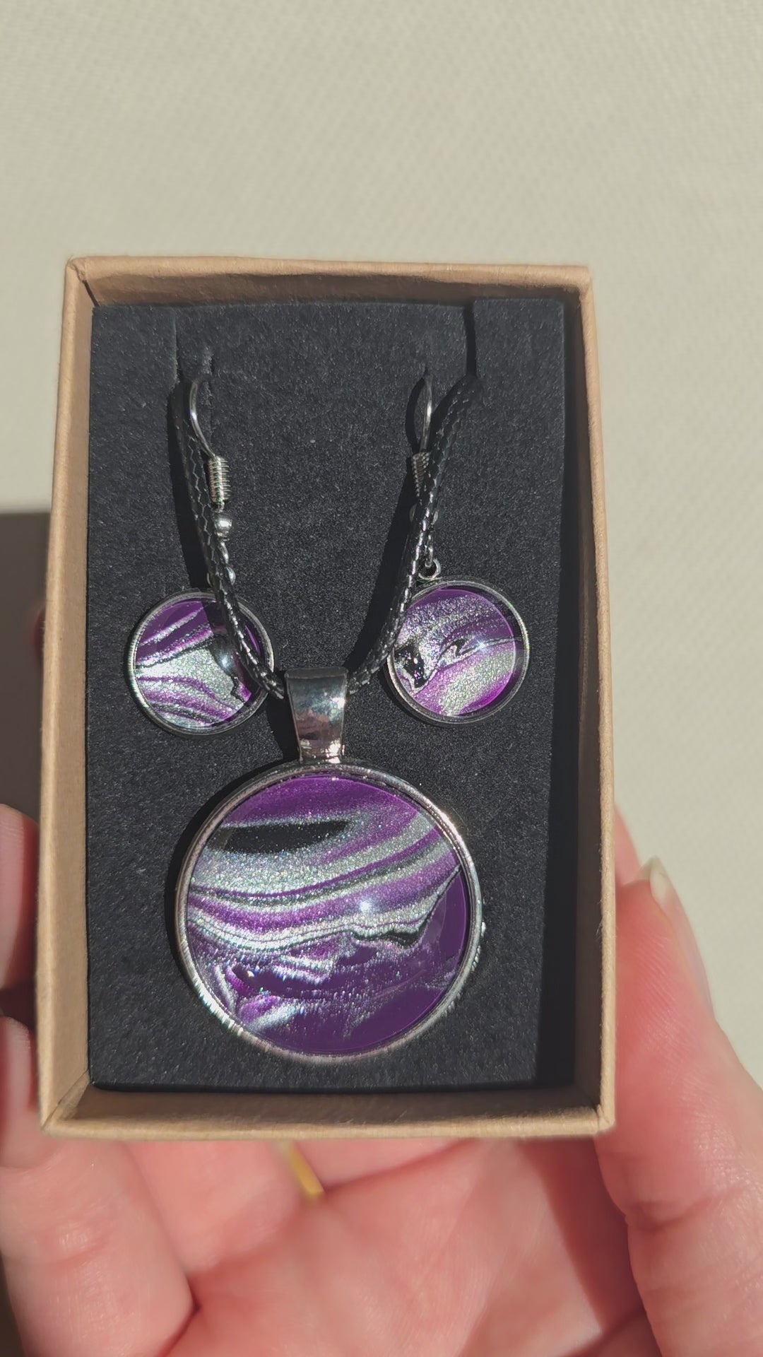 Cabochon-Schmuckset mit rundem Anhänger und zwei Ohrringen in dunklem Violett mit geschwungenen silbernen und irisierenden Perlmuttlinien sowie schwarzem Textilband.