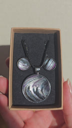 Cabochon-Schmuckset mit rundem Anhänger und zwei Ohrringen in Schwarz und Anthrazit mit geschwungenem silbernem Muster und violetten Akzenten.