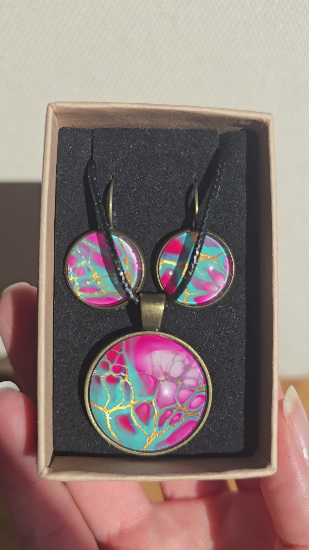Cabochon-Schmuckset mit rundem Anhänger und zwei Ohrringen in Türkis und kräftigem Pink mit verzweigten goldenen Linien und schwarzem Textilband in einer Schachtel.