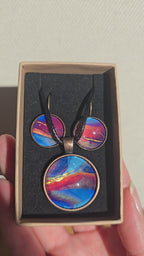 Rundes Cabochon-Schmuckset mit Anhänger und Ohrringen an schwarzem Band, mit abstraktem Muster in Blau, Cyan, Pink und Violett sowie feinen goldenen Linien.