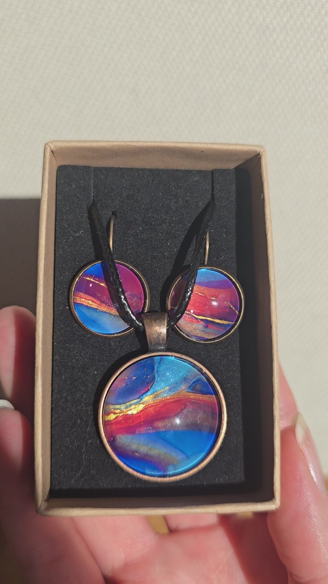 Rundes Cabochon-Schmuckset mit Anhänger und Ohrringen an schwarzem Band, mit abstraktem Muster in Blau, Cyan, Pink und Violett sowie feinen goldenen Linien.