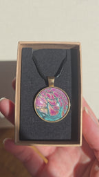 Runder Cabochon-Anhänger an schwarzem Band mit organischem, dunkel umrandetem Muster in Pink, Rosa, Türkis und warm schimmernden Nuancen.