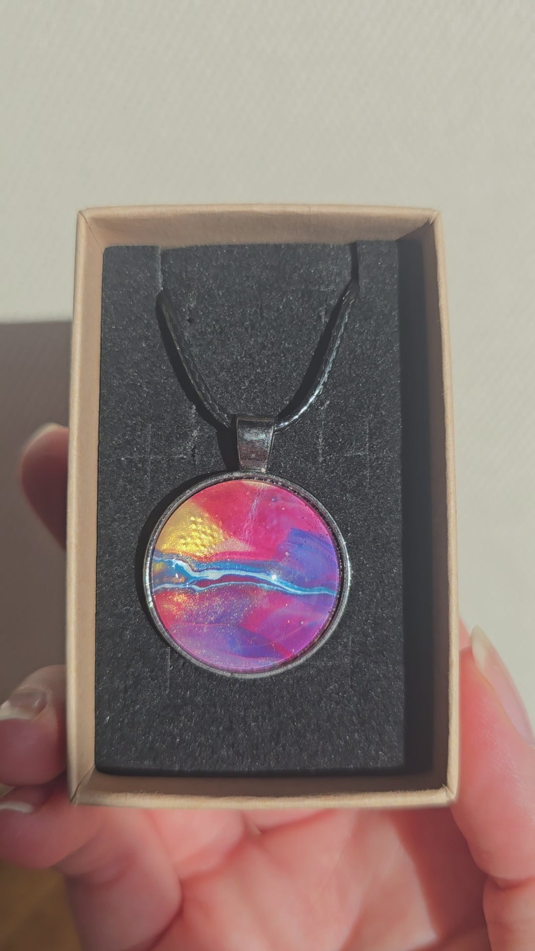 Runder Cabochon-Anhänger an schwarzem Band mit geschichtetem Muster in Fuchsia, Pink, Violett, Blau, Türkis und einem goldenen Akzent.