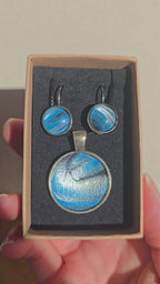 Rundes Cabochon-Schmuckset mit Anhänger und Ohrringen an schwarzem Band, mit fliessendem Muster in Cyan und Blau, silbrigem Perlmuttschimmer und dunklen Linien.