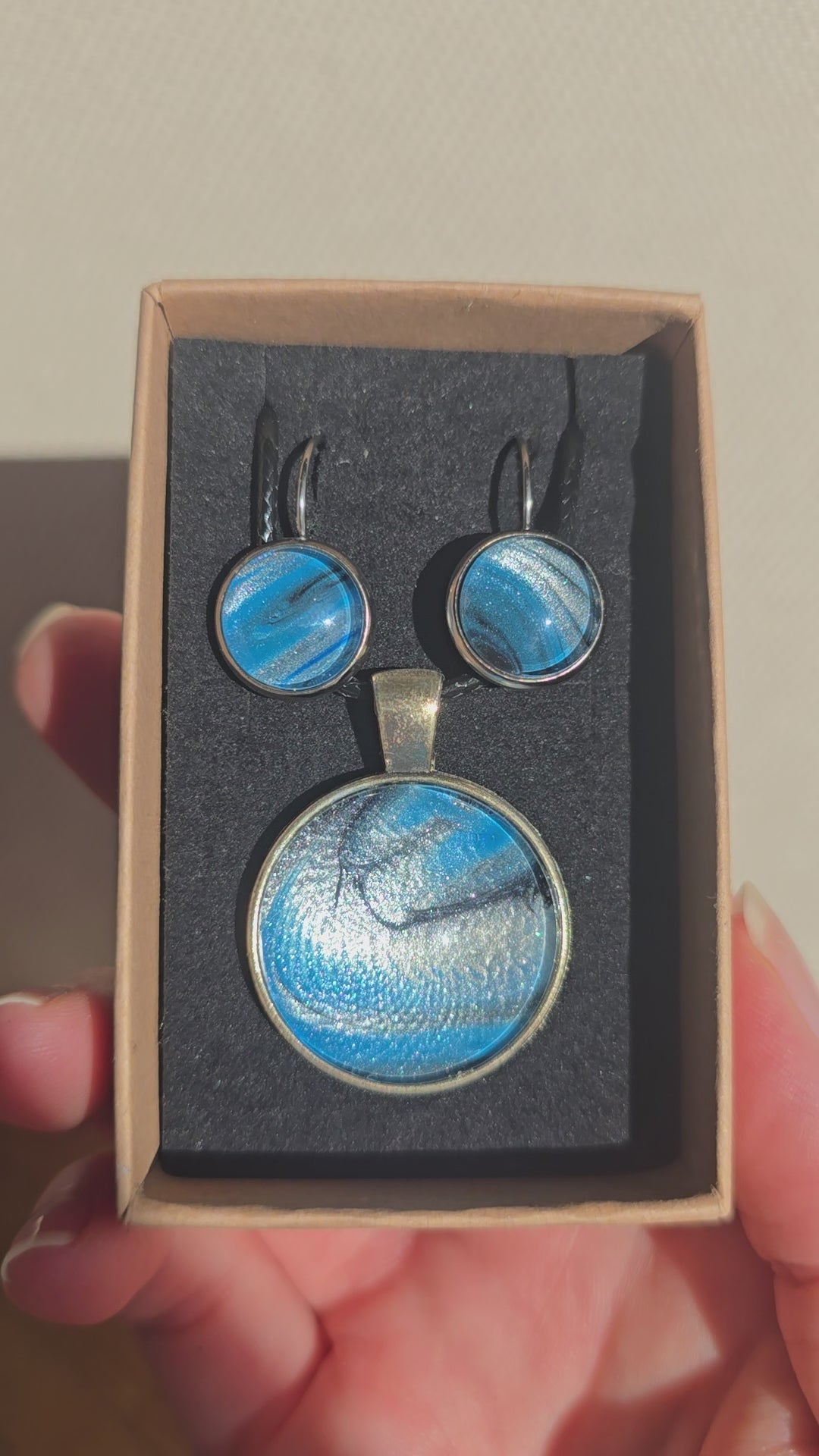 Rundes Cabochon-Schmuckset mit Anhänger und Ohrringen an schwarzem Band, mit fliessendem Muster in Cyan und Blau, silbrigem Perlmuttschimmer und dunklen Linien.