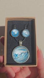 Rundes Cabochon-Schmuckset mit Anhänger und Ohrringen an schwarzem Band, mit fliessendem abstraktem Muster in Cyan, Blau, Grau und Schwarz.