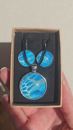 Rundes Cabochon-Schmuckset mit Anhänger und Ohrringen an schwarzem Band, mit organischem Muster in Türkis, Cyan und Blau sowie silbernem Schimmer.