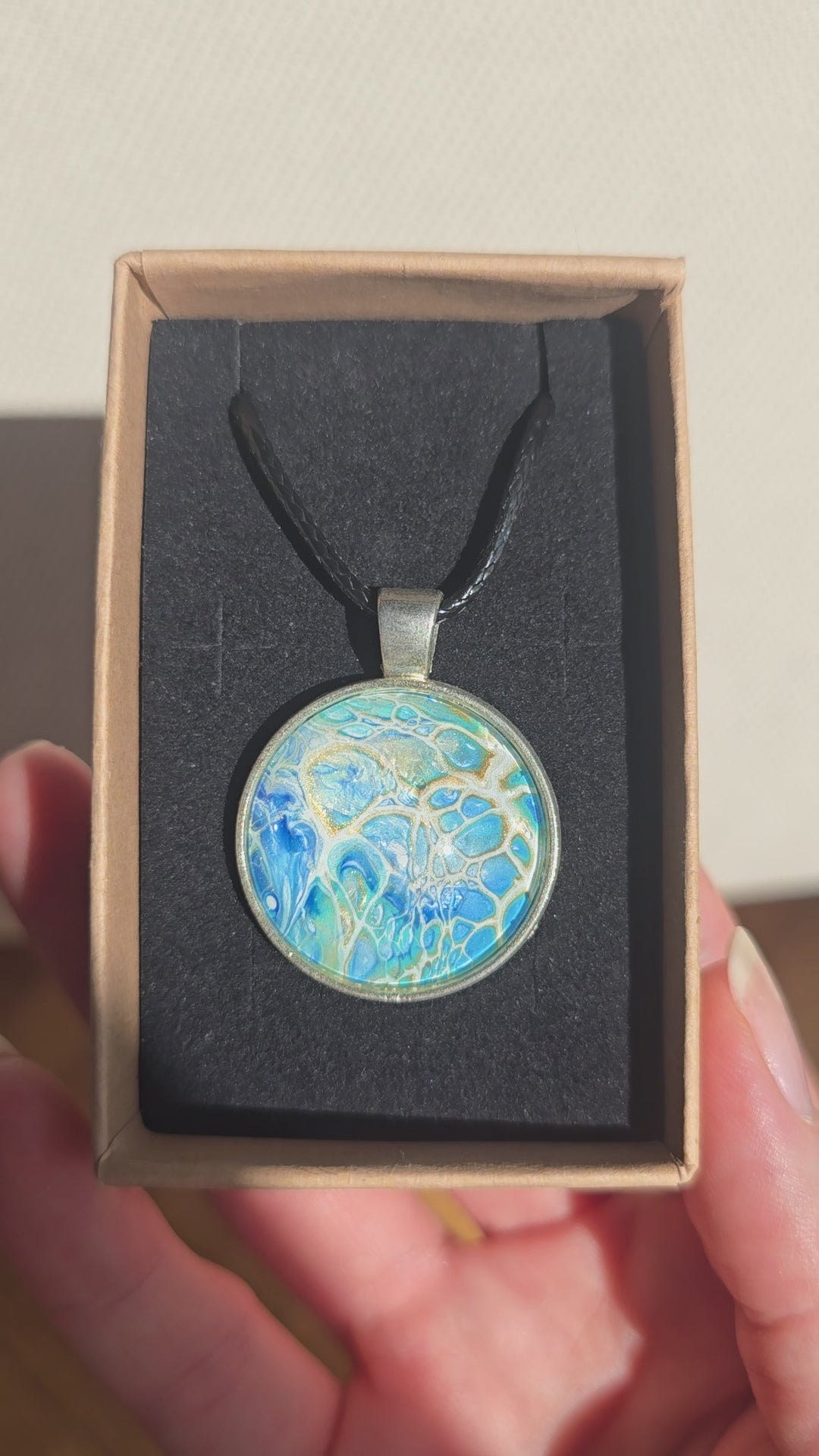Runder Cabochon-Anhänger an schwarzem Band mit filigranem, netzartigem Muster in Aqua-, Türkis-, Hellblau- und Cremetönen.