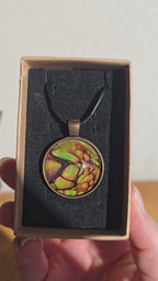 Runde Cabochon Kette Solas Forest Flame in Grün, Orange und Gold mit dunklen Strukturen, bronzefarbener Fassung und schwarzem Band.