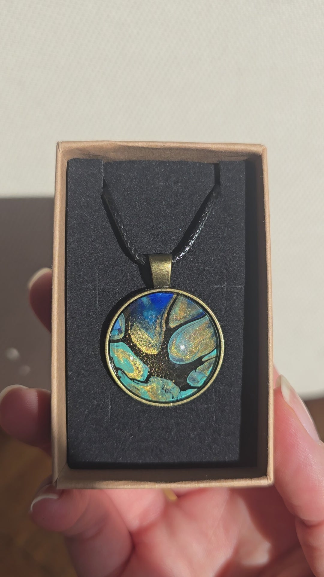 Runder Cabochon-Anhänger an schwarzem Band mit organischem Muster in Türkis, tiefem Blau, Schwarz und warm schimmernden Goldtönen.