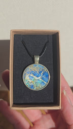 Runder Cabochon-Anhänger an schwarzem Band mit abstraktem, mineralischem Muster in Blau, Türkis, Weiss und Gold.