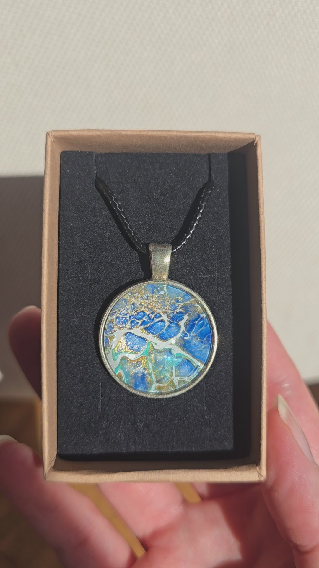 Runder Cabochon-Anhänger an schwarzem Band mit abstraktem, mineralischem Muster in Blau, Türkis, Weiss und Gold.