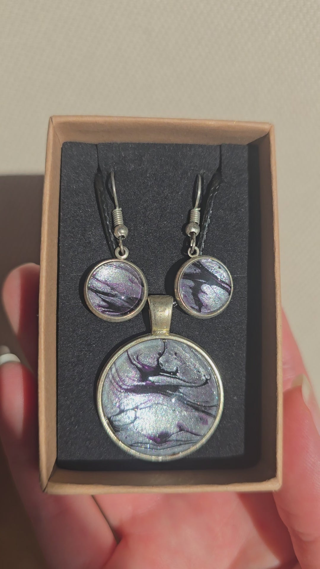 Rundes Cabochon-Schmuckset mit Anhänger und Ohrringen an schwarzem Band, mit silbrigem Perlmuttschimmer, Violett und fliessenden schwarzen Linien.