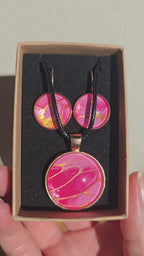 Cabochon-Schmuckset mit Anhänger und Ohrringen in Pink- und Rosatönen mit goldenen, fliessenden Linien und schwarzem Textilband in einer Schachtel.