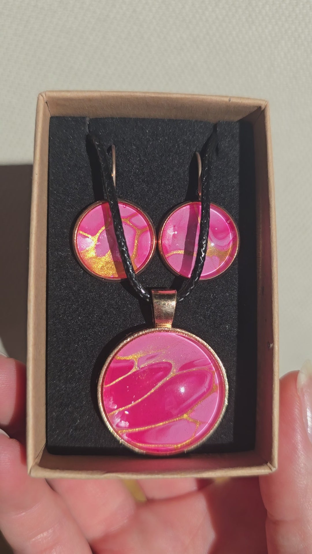 Cabochon-Schmuckset mit Anhänger und Ohrringen in Pink- und Rosatönen mit goldenen, fliessenden Linien und schwarzem Textilband in einer Schachtel.