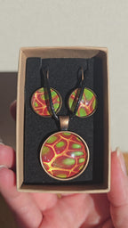 Cabochon-Schmuckset mit rundem Anhänger und zwei Ohrringen mit organischem Muster in Rostrot, Orange und leuchtendem Grün, kupferfarbenen Fassungen und schwarzem Textilband.