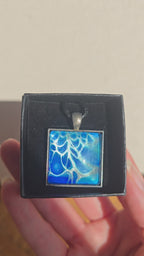 Quadratischer Cabochon-Anhänger an schwarzem Band mit abstraktem, fliessendem Muster in intensiven Blau-, Türkis- und hell schimmernden Nuancen.