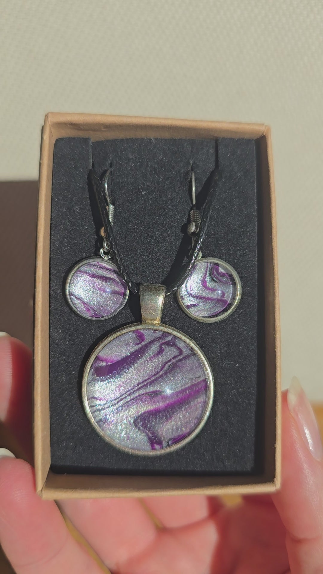 Cabochon-Schmuckset mit rundem Anhänger und zwei Ohrringen mit fliessendem Muster in Violett, Lila und schimmerndem Silber sowie schwarzem Textilband in einer Schachtel.