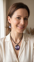Frau trägt Cabochon-Schmuckset mit rundem Anhänger und zwei Ohrringen in dunklem Violett mit geschwungenen silbernen und irisierenden Perlmuttlinien sowie schwarzem Textilband.