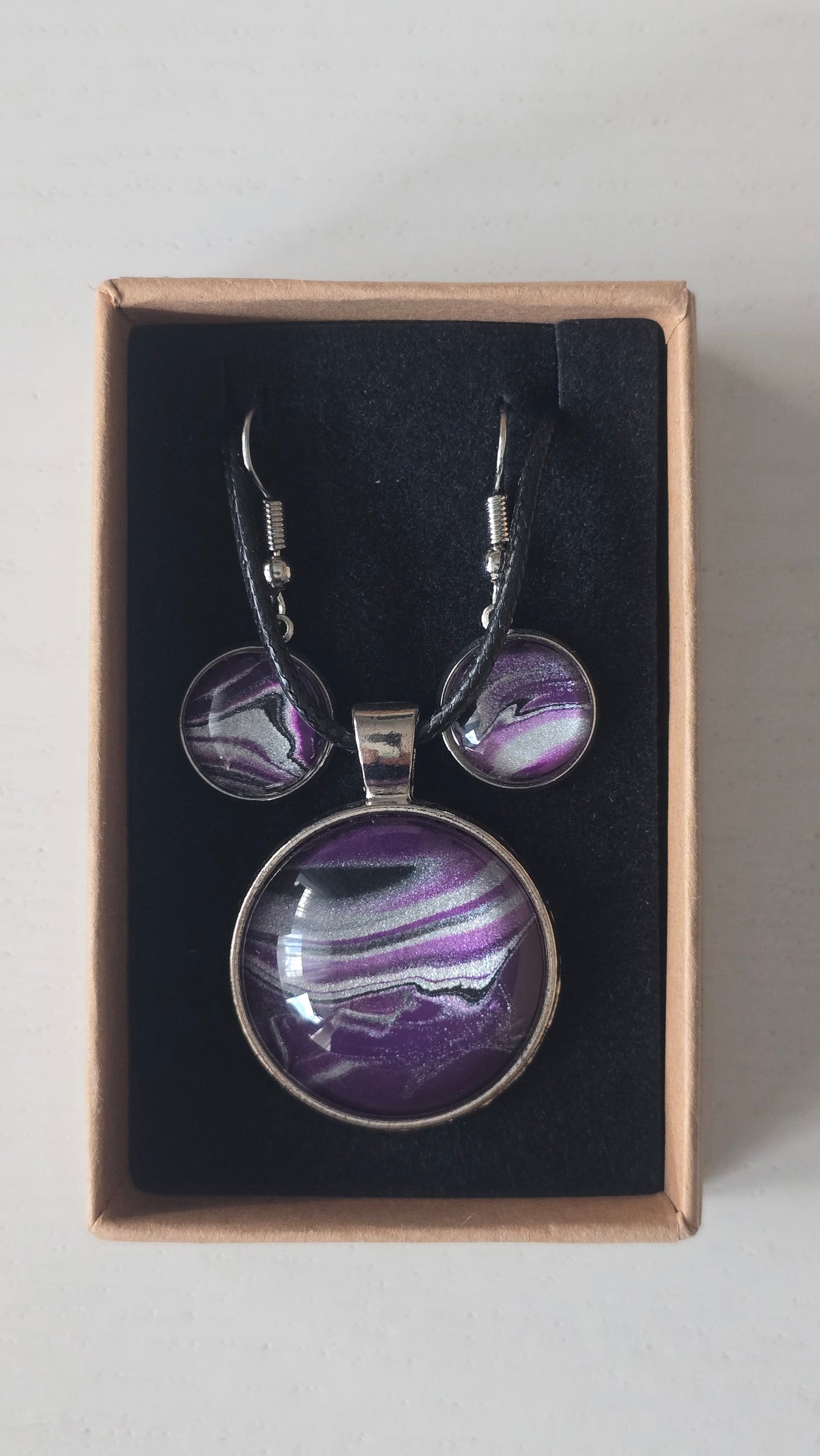 Cabochon-Schmuckset mit rundem Anhänger und zwei Ohrringen in dunklem Violett mit geschwungenen silbernen und irisierenden Perlmuttlinien sowie schwarzem Textilband.