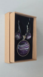 Cabochon-Schmuckset mit rundem Anhänger und zwei Ohrringen in dunklem Violett mit geschwungenen silbernen und irisierenden Perlmuttlinien sowie schwarzem Textilband.