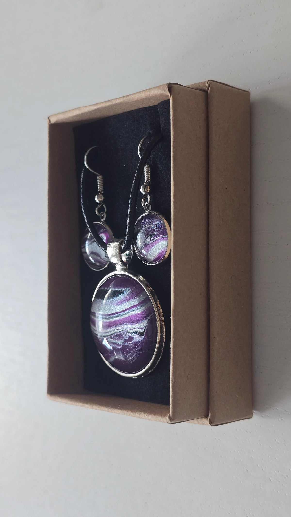 Cabochon-Schmuckset mit rundem Anhänger und zwei Ohrringen in dunklem Violett mit geschwungenen silbernen und irisierenden Perlmuttlinien sowie schwarzem Textilband.