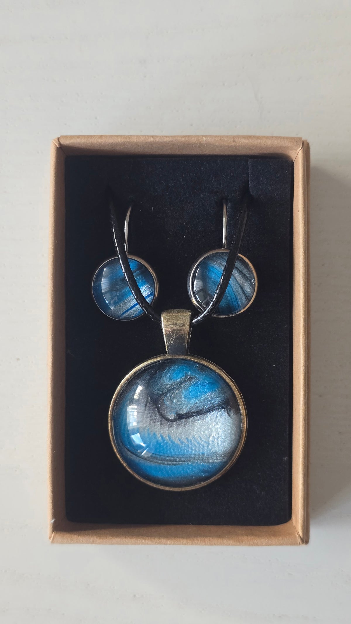 Rundes Cabochon-Schmuckset mit Anhänger und Ohrringen an schwarzem Band, mit fliessendem Muster in Cyan und Blau, silbrigem Perlmuttschimmer und dunklen Linien.