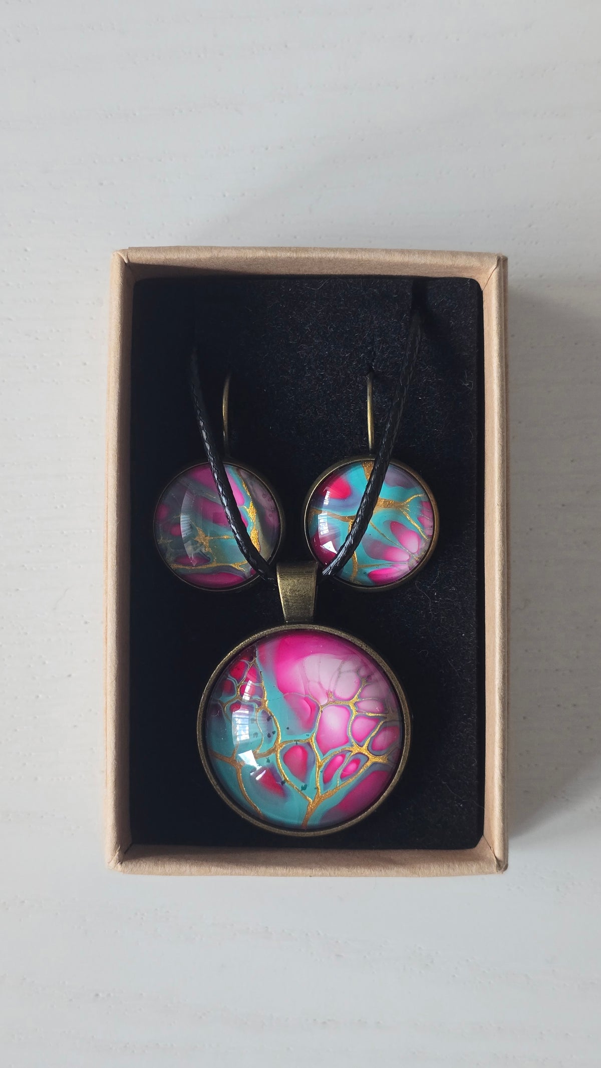 Cabochon-Schmuckset mit rundem Anhänger und zwei Ohrringen in Türkis und kräftigem Pink mit verzweigten goldenen Linien und schwarzem Textilband in einer Schachtel.