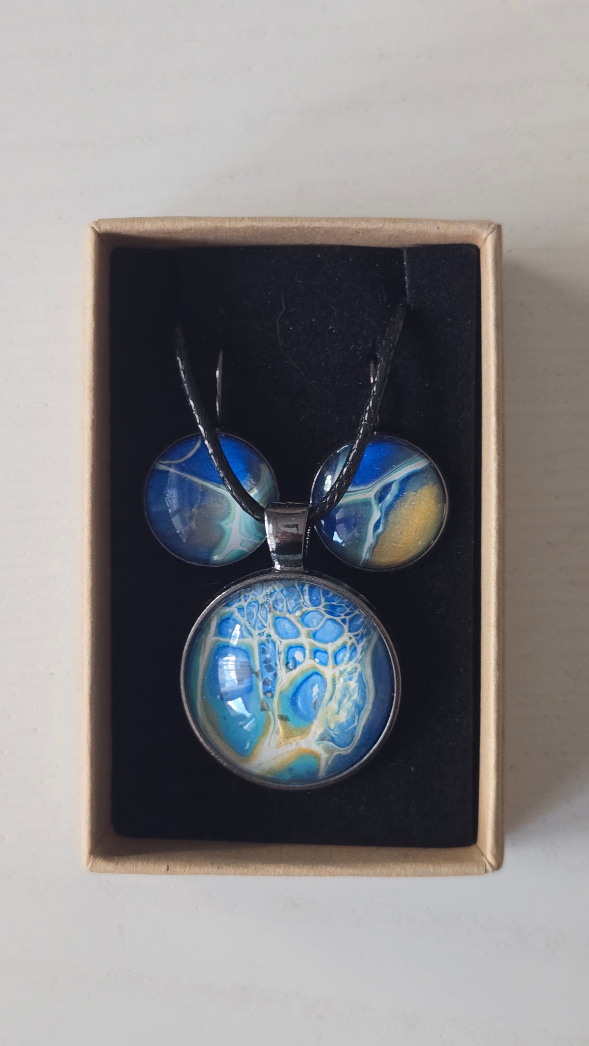 Rundes Cabochon Schmuckset mit schwarzer Kette und zwei Ohrringen, abstrakte Muster in Blau, Türkis, Weiss und Gold mit feinen netzartigen Strukturen.