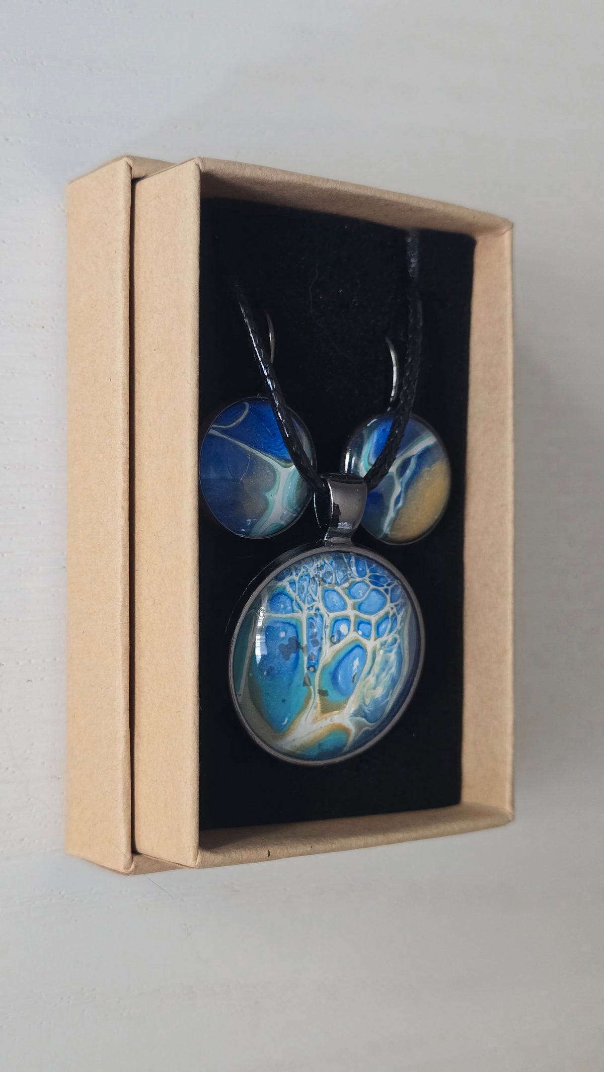 Rundes Cabochon Schmuckset mit schwarzer Kette und zwei Ohrringen, abstrakte Muster in Blau, Türkis, Weiss und Gold mit feinen netzartigen Strukturen.