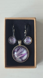 Cabochon-Schmuckset mit rundem Anhänger und zwei Ohrringen mit fliessendem Muster in Violett, Lila und schimmerndem Silber sowie schwarzem Textilband in einer Schachtel.