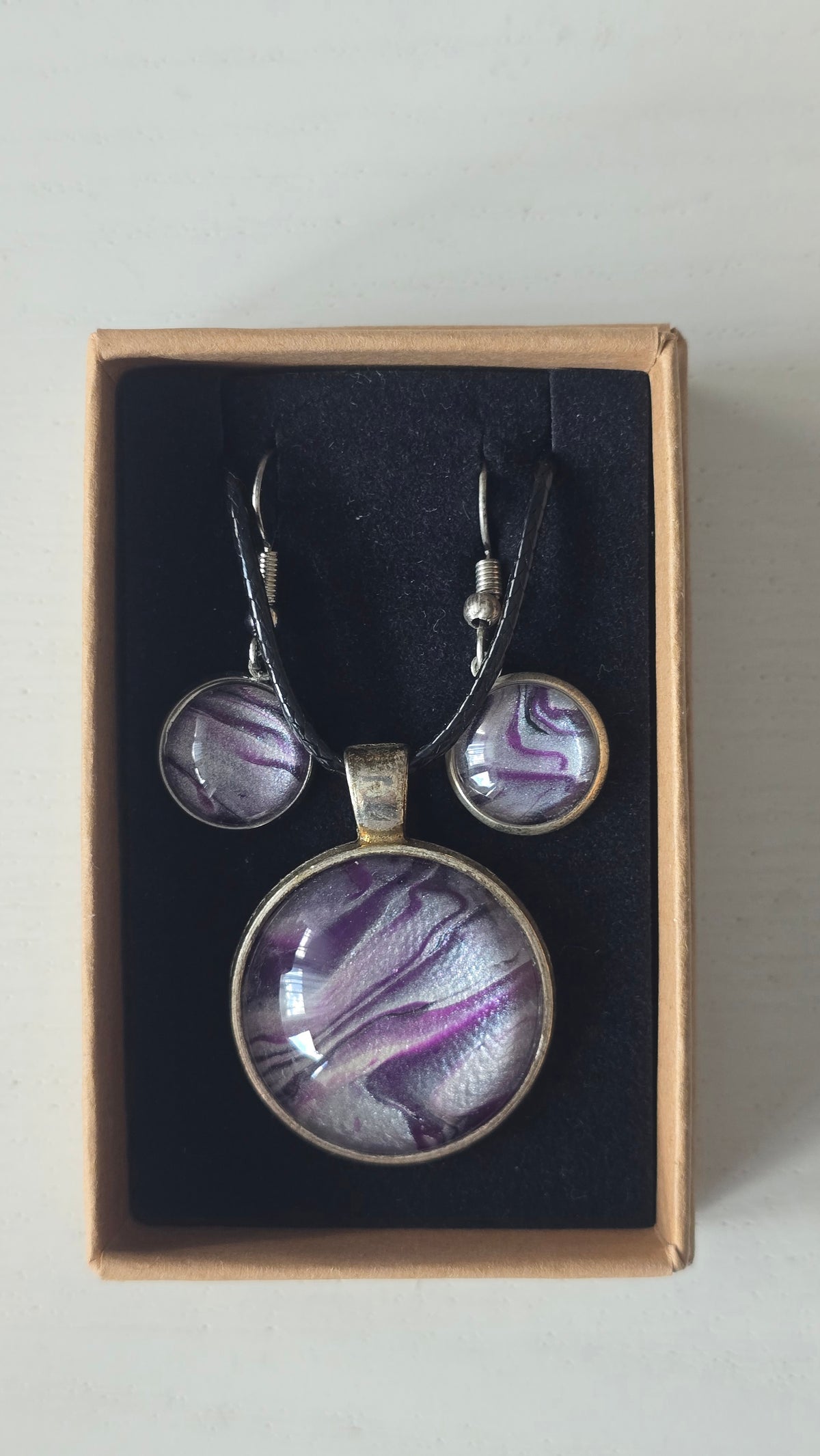 Cabochon-Schmuckset mit rundem Anhänger und zwei Ohrringen mit fliessendem Muster in Violett, Lila und schimmerndem Silber sowie schwarzem Textilband in einer Schachtel.