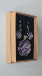 Cabochon-Schmuckset mit rundem Anhänger und zwei Ohrringen mit fliessendem Muster in Violett, Lila und schimmerndem Silber sowie schwarzem Textilband in einer Schachtel.