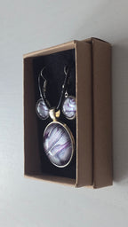 Cabochon-Schmuckset mit rundem Anhänger und zwei Ohrringen mit fliessendem Muster in Violett, Lila und schimmerndem Silber sowie schwarzem Textilband in einer Schachtel.