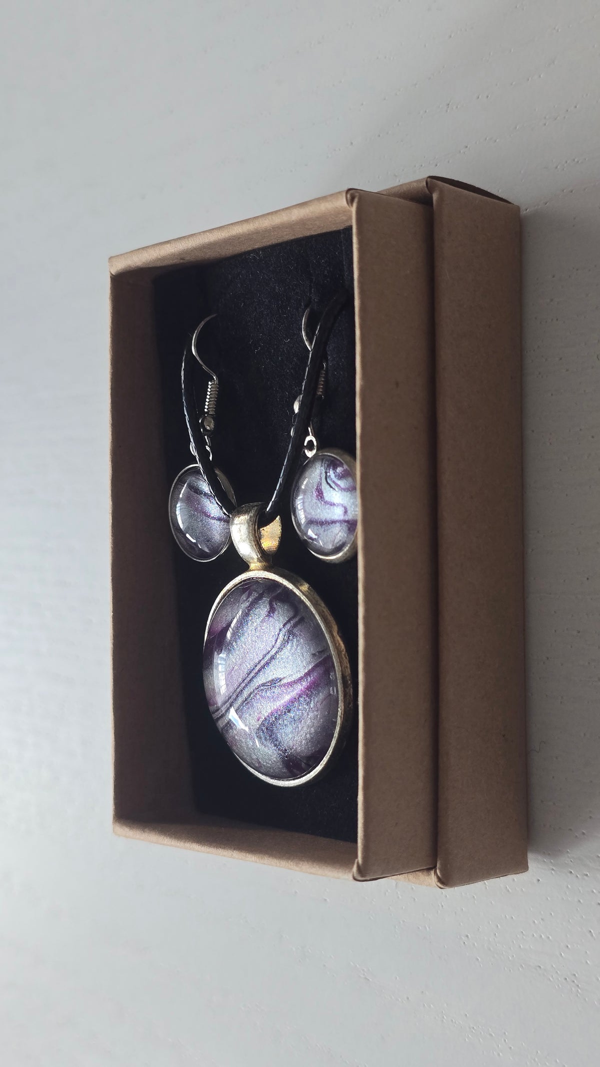 Cabochon-Schmuckset mit rundem Anhänger und zwei Ohrringen mit fliessendem Muster in Violett, Lila und schimmerndem Silber sowie schwarzem Textilband in einer Schachtel.