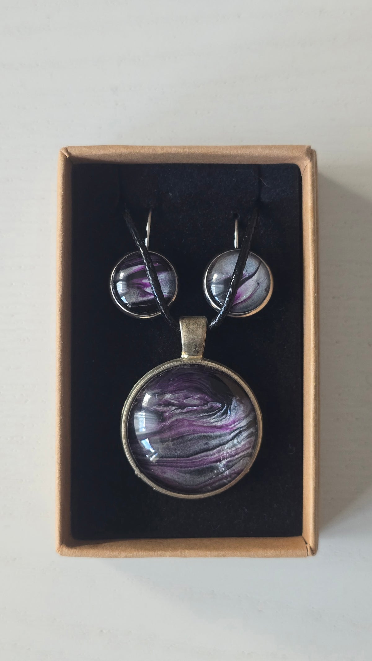 Rundes Cabochon-Schmuckset mit Anhänger und Ohrringen an schwarzem Band, mit fliessendem Muster in Violett, Pflaume, Silber und Schwarz.