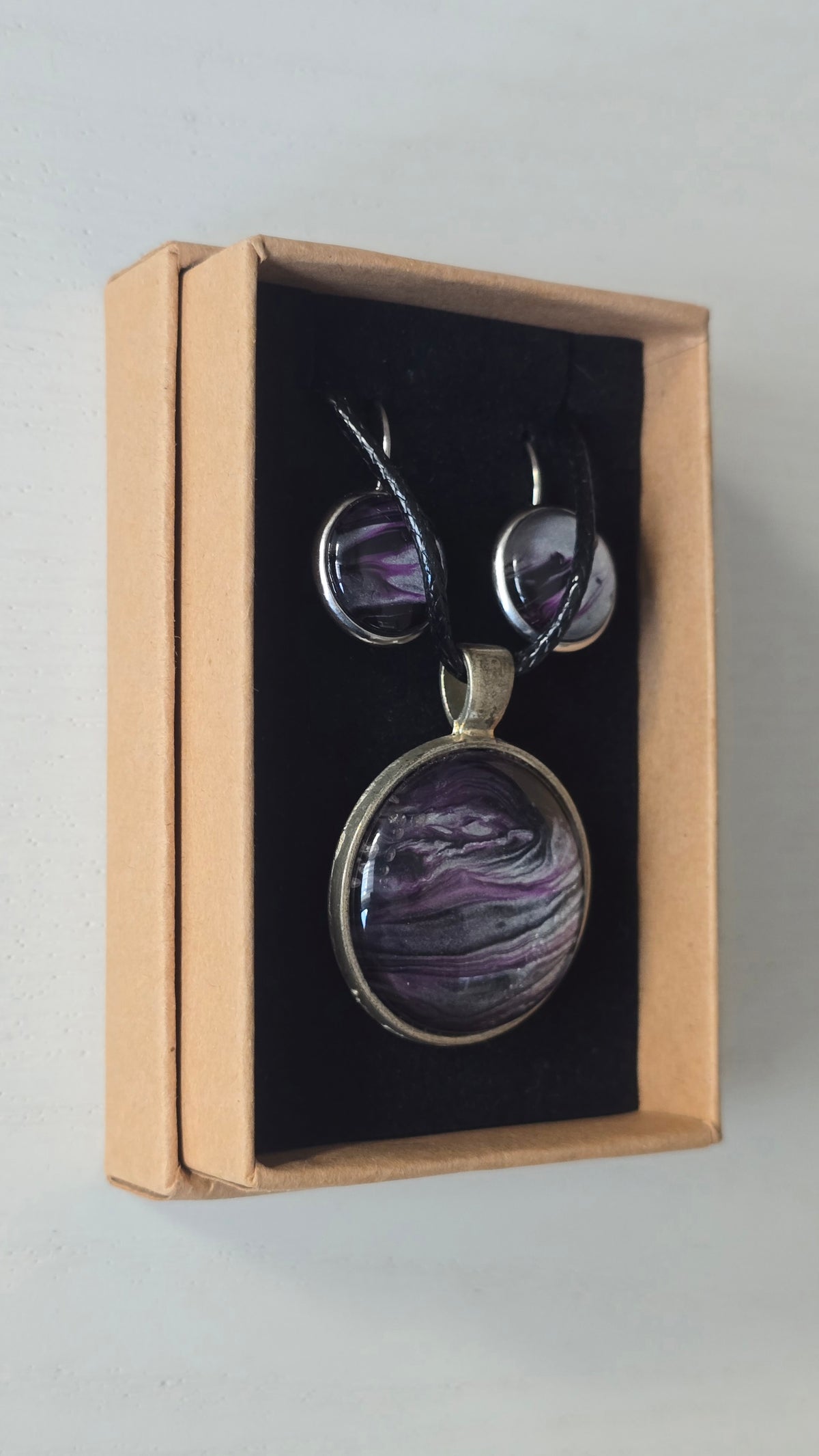 Rundes Cabochon-Schmuckset mit Anhänger und Ohrringen an schwarzem Band, mit fliessendem Muster in Violett, Pflaume, Silber und Schwarz.